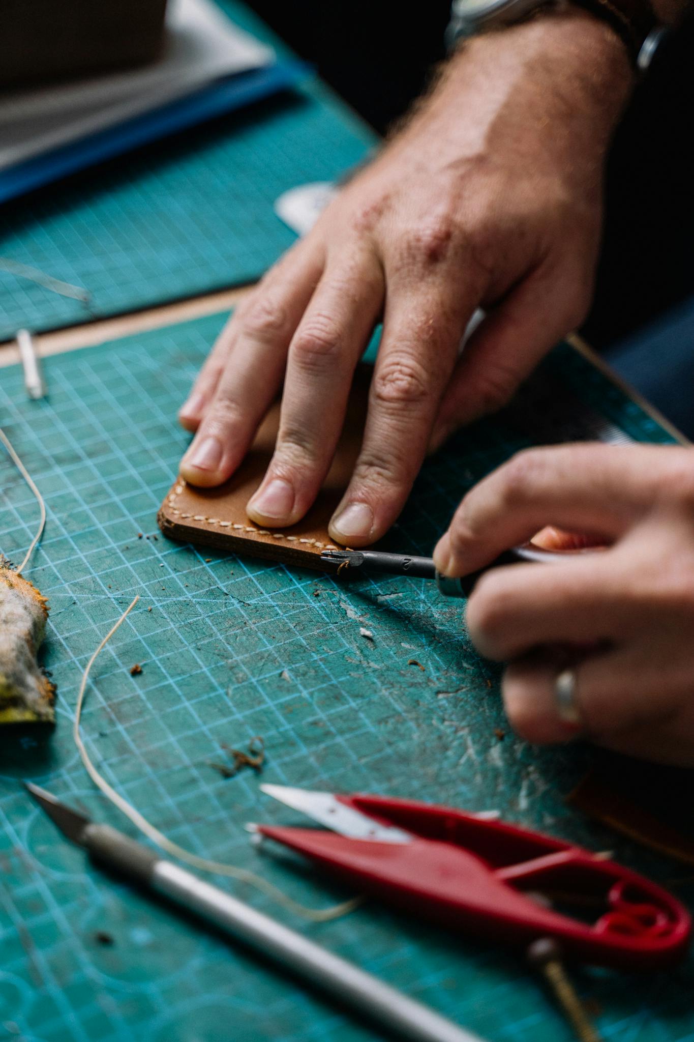 Hands working with leather and tools on a green cutting mat in a workshop setting.